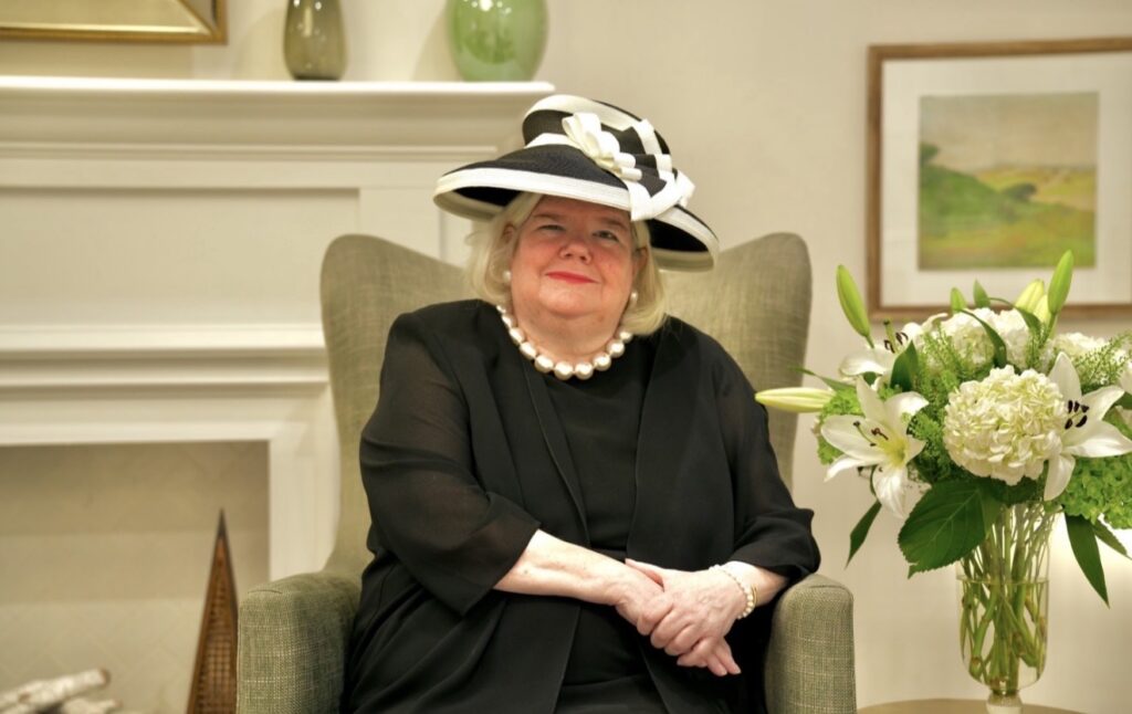 Michelle Leonard, Main Line REALTOR®, wearing a black-and-white hat and seated beside white flowers.
